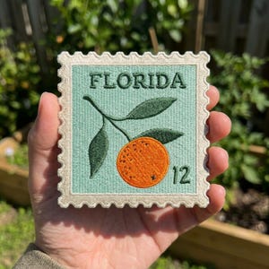 May include: A square embroidered patch shaped like a postage stamp with a light green background. It features a bright orange citrus fruit with dark green leaves and the text FLORIDA and the number 12 in dark green thread with a cream border.