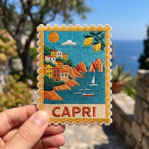 May include: A rectangular embroidered patch shaped like a postage stamp featuring a coastal scene of Capri. The design includes colorful houses on a cliff, a bright sun, two yellow lemons, a sailboat, and the word CAPRI in orange letters at the bottom.