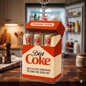 May include: A Diet Coke-themed keychain resembling a cigarette box, with the words "FRIDGE CIGS" on top. Inside are three miniature Diet Coke cans. The text "Diet Coke" and "JUST A LITTLE SOMETHING TO TAKE THE EDGE OFF" are also visible.