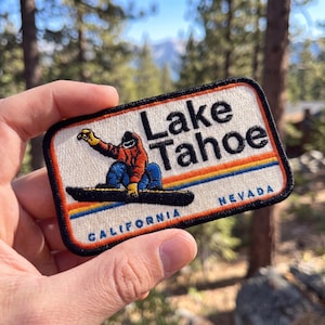 May include: A rectangular embroidered patch with a cream background and black border. It features a snowboarder in an orange jacket and blue pants, with the text Lake Tahoe in black, and California and Nevada in blue, plus a rainbow stripe.
