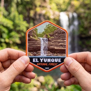May include: A hexagonal embroidered patch featuring a landscape of a waterfall cascading over brown layered rock cliffs with green foliage. The bottom black border displays the white text EL YUNQUE, orange text NATIONAL FOREST, and 1903.
