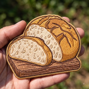 May include: An embroidered patch featuring a golden-brown loaf of bread sliced into two pieces, resting on a dark brown wooden cutting board. The bread has a textured crust and a light cream-colored interior with visible grain details in brown thread.