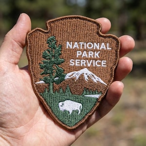 May include: A shield-shaped embroidered patch with a brown border and background. It features a green pine tree, a white mountain peak, and a white bison silhouette. The text NATIONAL PARK SERVICE is embroidered in white capital letters.