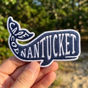 May include: A dark navy blue embroidered patch shaped like a whale, featuring a white border and white stitched text that reads CAPE COD on the tail and NANTUCKET across the body of the whale, with a textured fabric finish throughout the design.
