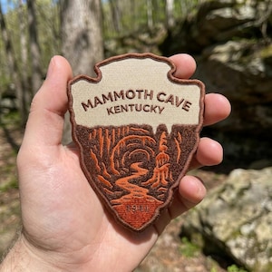 May include: A shield-shaped embroidered patch featuring a beige upper section with the text MAMMOTH CAVE KENTUCKY in brown. The lower section depicts a brown and orange cave landscape with a winding path and the year 1941 at the bottom edge.