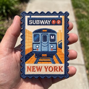 May include: A rectangular embroidered patch shaped like a postage stamp with a dark blue border. The design features a blue subway train on tracks, a sign reading SUBWAY 1 2, and the text NEW YORK in orange at the bottom on a cream background.