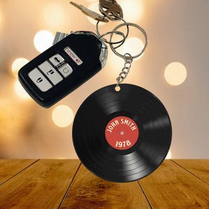 May include: A black car key fob and a keychain with a black vinyl record design. The record has a red center with the name "JOHN SMITH" and the year "1978" printed on it. The keychain is attached to a silver key ring.