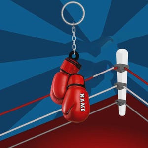 May include: A red boxing glove keychain with the word "NAME" printed on one glove. The keychain hangs in front of a boxing ring with red and white ropes, and a blue background.