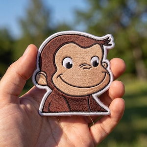 May include: An embroidered patch featuring the face of a brown monkey with a light tan face, large black eyes, and a subtle smile. The design is outlined in a thick white border with detailed stitching throughout the brown and tan thread.
