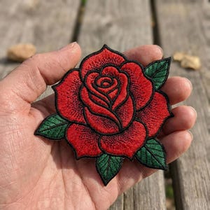 May include: An embroidered patch featuring a detailed red rose with layered petals and dark green leaves. The design is outlined in black thread, creating a clear contrast against the vibrant red and green colors of the floral embroidery.