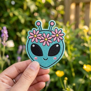May include: A teal alien patch with large black eyes, pink cheeks, and a smiling mouth. The alien wears a floral crown of pink flowers with yellow centers. The patch is embroidered with black thread and has a black border.