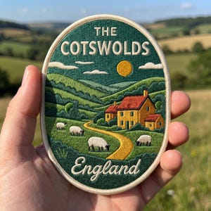 May include: An oval embroidered patch featuring a landscape of The Cotswolds, England. The design shows green rolling hills, a yellow sun, a yellow house with a red roof, and four white sheep grazing near a winding yellow path.