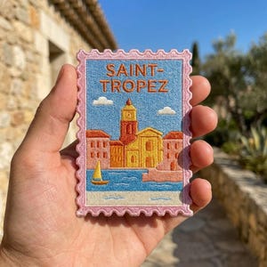 May include: A rectangular embroidered patch with a scalloped pink border featuring a Saint-Tropez town scene. The design includes a central bell tower, yellow and orange buildings, a blue sea with a yellow sailboat, and two white clouds.