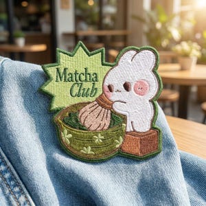 May include: An embroidered patch featuring a white cartoon rabbit holding a bamboo whisk in a green matcha bowl. The patch has a light green starburst background with the text "Matcha Club" in dark green lettering, all stitched onto blue denim fabric.