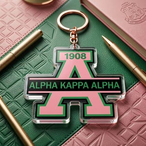 May include: A clear acrylic keychain with a rose gold keyring. The keychain features the text "ALPHA KAPPA ALPHA" in black and green, with the number "1908" in green. The letter "A" is pink with green trim.