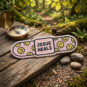 May include: A pink adhesive bandage patch featuring a repeating pattern of yellow smiley faces and a central square with the black embroidered text JESUS HEALS, positioned next to a brass compass on a weathered wooden surface.