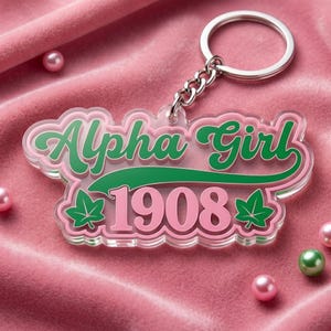 May include: A clear acrylic keychain with the words "Alpha Girl 1908" in green and pink script. The keychain has a silver chain and ring, and is decorated with green ivy leaves. The background is a pink velvet surface.