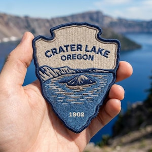 May include: A shield-shaped embroidered patch featuring a tan sky and dark blue water depicting Crater Lake, Oregon. The text CRATER LAKE OREGON is stitched in dark blue, with the year 1902 at the bottom. The patch has a dark blue border.