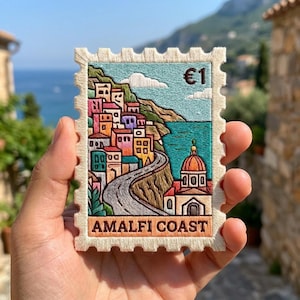 May include: A rectangular embroidered patch shaped like a postage stamp featuring a colorful coastal scene of the Amalfi Coast. The design includes stacked buildings, a winding road, a domed church, the text €1, and the words AMALFI COAST.