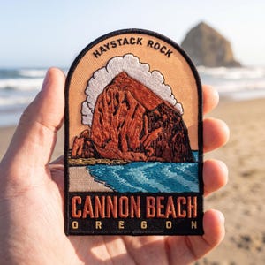May include: An embroidered patch featuring a depiction of Haystack Rock in Cannon Beach, Oregon. The design includes a large brown rock formation, white clouds, and blue water, with the text HAYSTACK ROCK and CANNON BEACH OREGON.