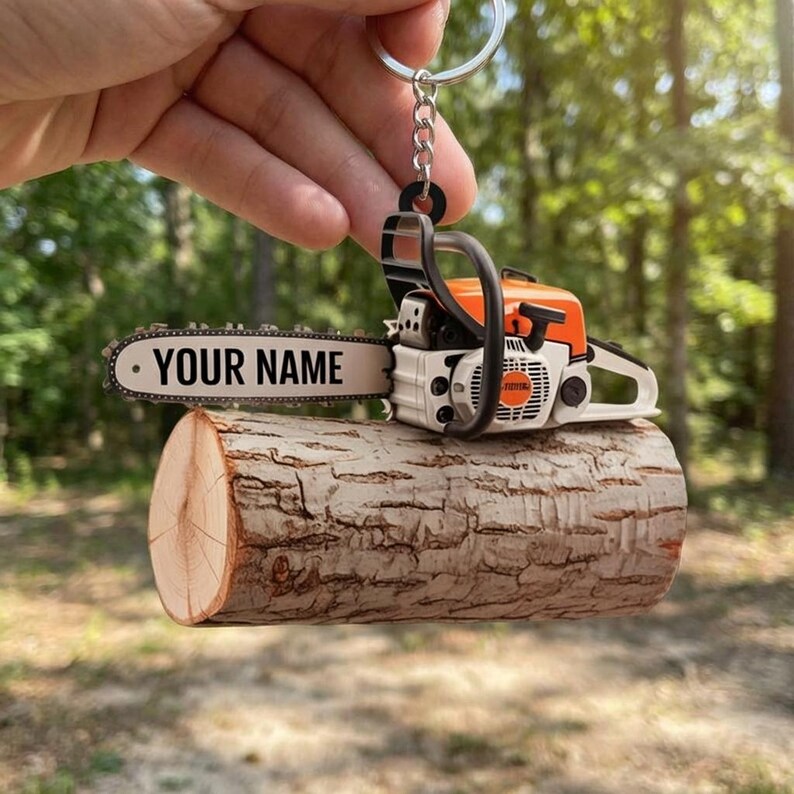 May include: A miniature chainsaw keychain with an orange and white design, resting on a small wooden log. The chainsaw blade has the text "YOUR NAME" printed on it. The keychain is held up by a silver keyring.