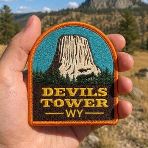 May include: An embroidered patch featuring a light-colored rock formation against a bright blue sky, set above a dark green forest. The bottom section is dark brown with the yellow embroidered text "DEVILS TOWER WY" and a horizontal line.