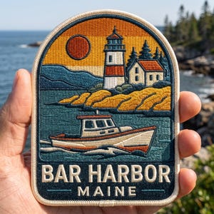 May include: An embroidered patch featuring a coastal scene with a lighthouse, a small boat on the water, and a sunset. The design includes a white lighthouse with a red stripe, a house, trees, and the text BAR HARBOR MAINE in white lettering.