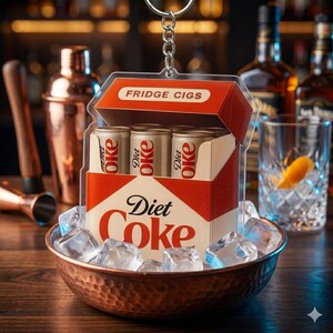 May include: A quirky keychain shaped like a Diet Coke box, complete with three miniature cans. The box is red and white, labelled "Fridge Cigs". It sits in a copper bowl filled with ice cubes. The background suggests a bar setting.