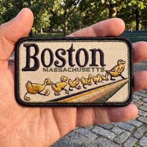 May include: A rectangular embroidered patch with a beige background and a black border. The text reads Boston MASSACHUSETTS in black lettering. Below the text, a line of yellow ducklings follows a larger yellow duck across a diagonal path.