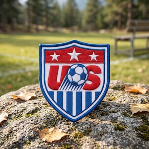 May include: A shield-shaped embroidered patch featuring the letters US in red with a blue and white soccer ball replacing the letter S. The top section is red with three white stars, and the bottom section has vertical blue and white stripes.