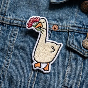 May include: Embroidered patch featuring a goose with a flower bouquet in its beak. The goose is cream-colored with orange feet and beak, and the number "3" is on its side. The patch is outlined in black and set against a denim jacket.