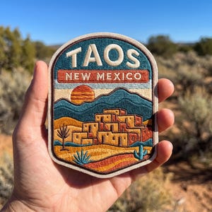 May include: An embroidered patch featuring a desert landscape with adobe buildings, a setting sun, and cacti. The text TAOS is stitched in cream above a red banner with the text NEW MEXICO. The design uses shades of blue, orange, and tan.