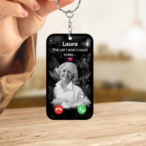 May include: A rectangular keychain with a silver keyring. The keychain features a black and white image of a woman and the text "Laura, The call I wish I could make..." with a red and green phone icon.
