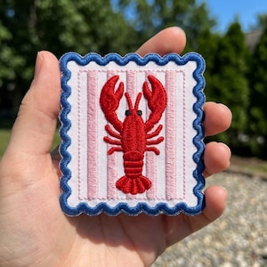 May include: A rectangular embroidered patch featuring a bright red lobster centered on a white background with vertical light pink stripes. The patch has a scalloped edge finished with a thick, dark blue embroidered border.