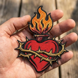 May include: An embroidered patch featuring a red heart encircled by a gold crown of thorns. Above the heart sits a dark vessel containing vibrant orange and yellow flames. The entire design is outlined in black thread with detailed stitching.