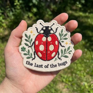 May include: An embroidered patch on a cream-colored fabric base featuring a red ladybug with cream spots, black legs, and antennae. Green leafy sprigs flank the insect, with small yellow stars and the text "the last of the bugs" in black.