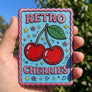 May include: A rectangular embroidered patch with a scalloped pink border on a light blue background. The text RETRO CHERRIES is stitched in pink. Two red cherries with a green leaf are centered, surrounded by small yellow stars and purple flowers.