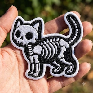 May include: An embroidered patch featuring a black cat silhouette with a white skeletal design. The cat is depicted in profile with a skull-shaped head, visible rib cage, spine, and tail bones, all outlined in white thread against a black background.