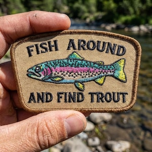 May include: A rectangular embroidered patch with a tan background and a dark brown stitched border. The patch features a colorful trout fish in the center with blue, pink, and yellow details. The text reads FISH AROUND AND FIND TROUT in black.