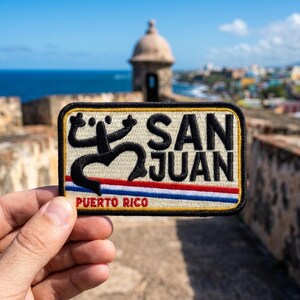 May include: A rectangular embroidered patch featuring a black Taino coqui symbol on the left, the text SAN JUAN in black, and a horizontal red, white, and blue stripe pattern above the red text PUERTO RICO on a cream-colored background.