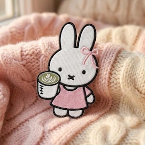 May include: An embroidered patch featuring a white rabbit character with long ears, a small pink bow, and a pink dress. The rabbit is holding a white mug containing a green beverage with latte art. The patch has a black outline and white stitching.
