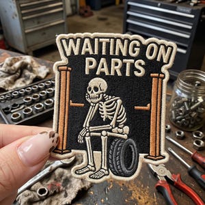 May include: Embroidered patch with the text "WAITING ON PARTS" above a skeleton sitting on a tire. The patch has a black background with tan and white accents. Tools and hardware are visible in the background.