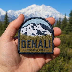 May include: A semi-circular embroidered patch featuring a mountain landscape with snow-capped peaks against a light blue sky, a dark green forest line, and the text DENALI NATIONAL PARK in dark blue lettering on a gold background.