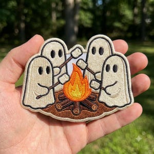 May include: An embroidered patch featuring four off-white ghosts sitting around a campfire. The ghosts are holding sticks with white marshmallows over a bright orange and yellow flame. The campfire is built on a base of brown logs and dirt.