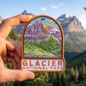 May include: An embroidered patch featuring a mountain landscape with purple peaks, green slopes, and a blue sky. The text 1910 is at the top, and the text GLACIER NATIONAL PARK is in orange letters on a white background at the bottom with an orange border.