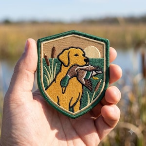 May include: A shield-shaped embroidered patch featuring a yellow Labrador retriever holding a mallard duck in its mouth. The scene includes green marsh grass, brown cattails, and a pale sun against a tan background, all bordered by a dark green edge.