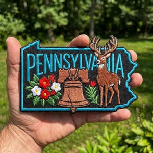 May include: A black embroidered patch shaped like the state of Pennsylvania with a bright blue border. It features the text PENNSYLVANIA in blue, a brown Liberty Bell, a brown deer with antlers, and red and white flowers with green leaves.