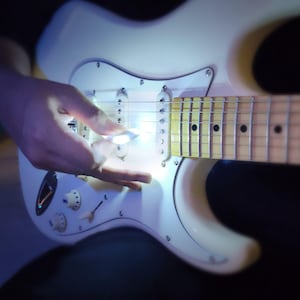 May include: A white electric guitar with a pick held over the strings. The guitar has a white pickguard, three pickups, and a maple neck. The image is well-lit, highlighting the details of the instrument.