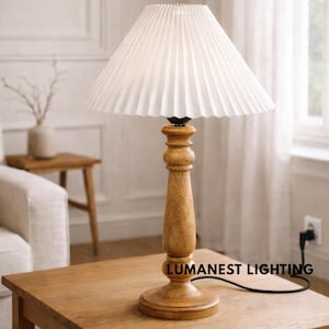 Puede incluir: Una lámpara de mesa de madera con una pantalla plisada blanca. La lámpara tiene una base de madera torneada y un cable negro. La lámpara está sobre una mesa de madera. La marca "LumaNest Lighting" es visible.