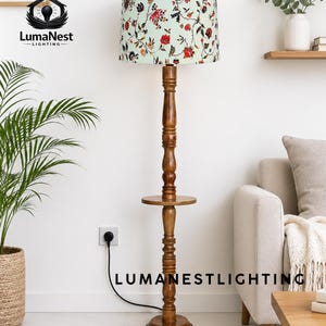 Könnte beinhalten: Eine hölzerne Stehlampe mit floral gemustertem Lampenschirm. Die Lampe hat einen braunen, gedrechselten Holzfuß und ein kleines rundes Regal. Der Lampenschirm hat einen hellgrünen Hintergrund mit roten und weißen Blumen und grünen Blättern. Der Text "LumaNestLighting" ist sichtbar.